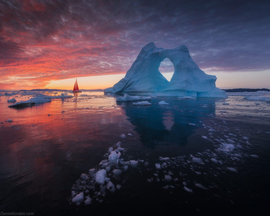 Red sailboat and giant arched iceberg at sunset in Greenland